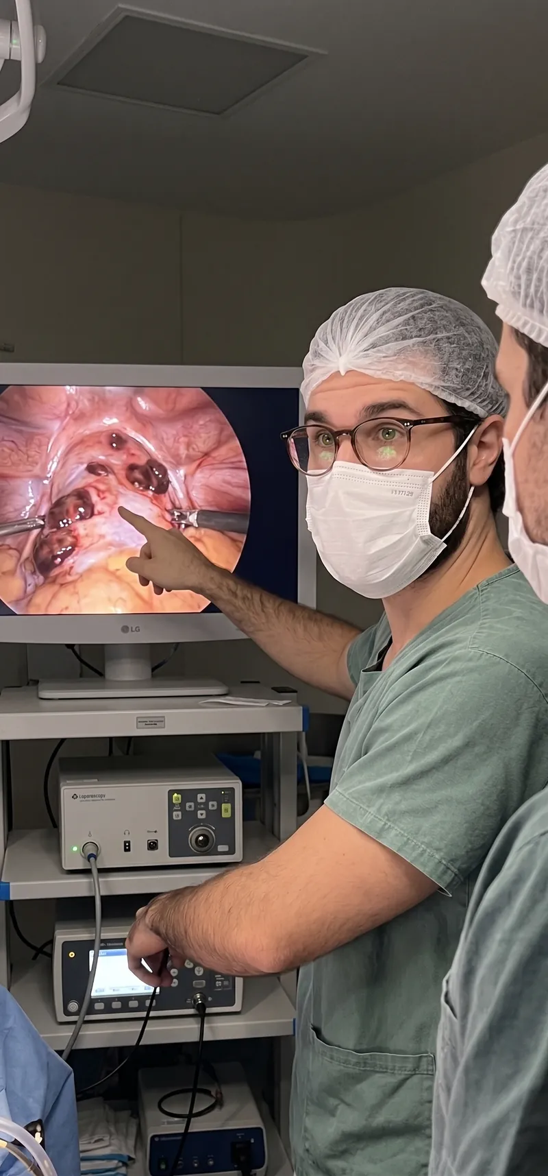 Dr. José Ricardo Ristow de touca, máscara e óculos, apontando para o monitor de vídeo da laparoscopia no centro cirúrgico