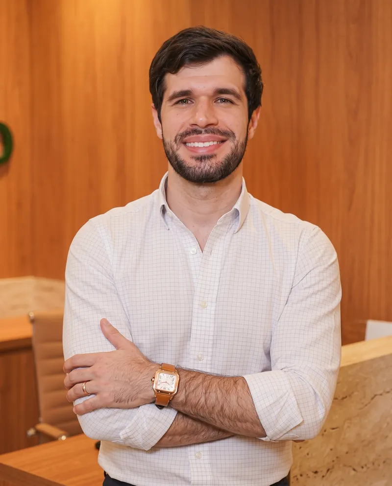 Dr. José Ricardo Ristow sorrindo de braços cruzados no consultório, diante de uma parede de madeira