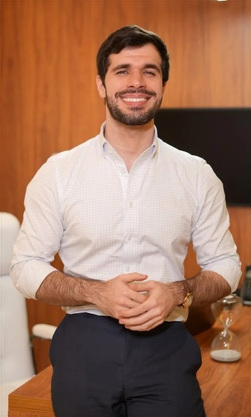 Dr. José Ricardo Ristow sorrindo de mãos entrelaçadas, apoiado na mesa do consultório