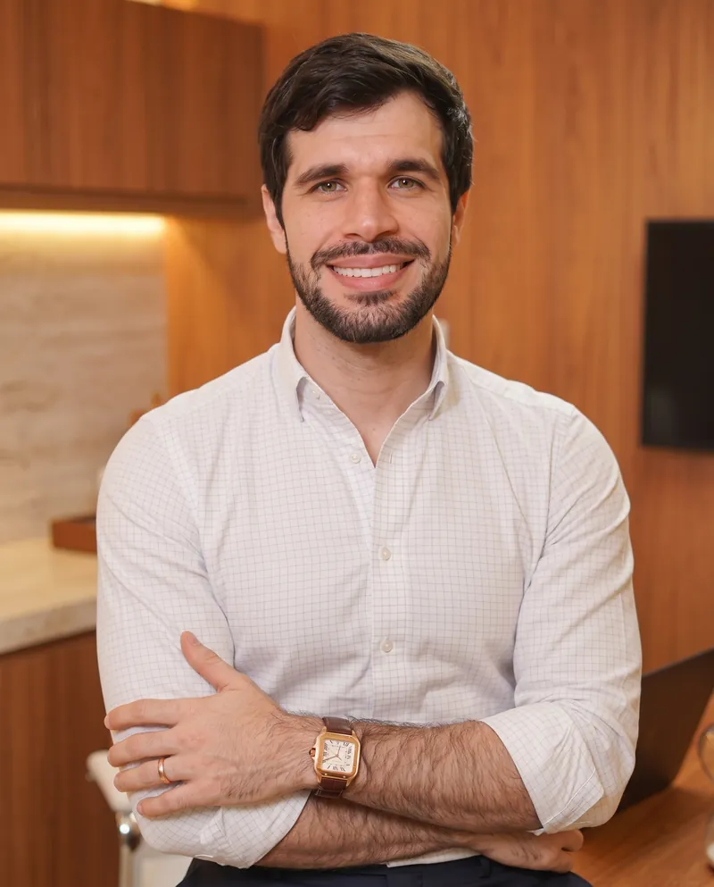 Dr. José Ricardo Ristow sorrindo de braços cruzados, apoiado na mesa do consultório
