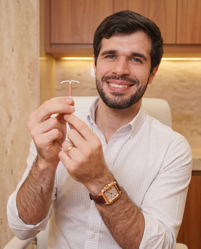 Dr. José Ricardo Ristow sorrindo no consultório, segurando um DIU em forma de T com as duas mãos