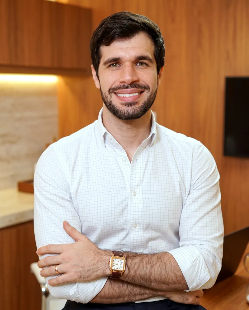 Dr. José Ricardo Ristow, ginecologista, sorrindo de braços cruzados no consultório, de camisa branca